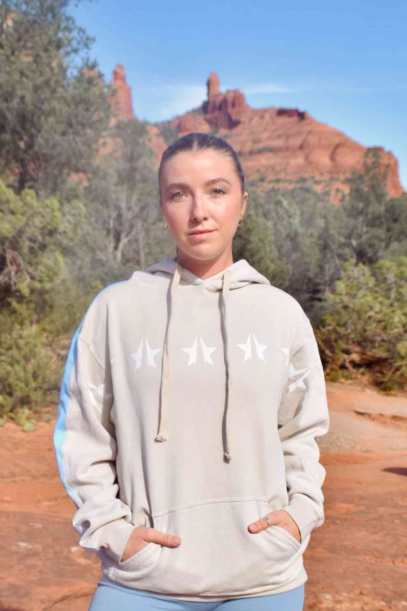 Person wearing a GFLApparel GFL Stars Hoodie in Dune stands outdoors with red rock formations and greenery in the background.