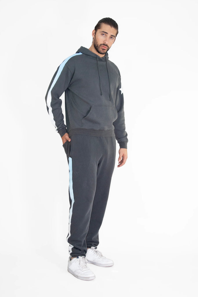 A man wearing a dark grey Light Worker Hoodie in Space Glow from GFLApparel and matching sweatpants with white and light blue stripes stands facing the camera, hands in pockets, against a plain white background.
