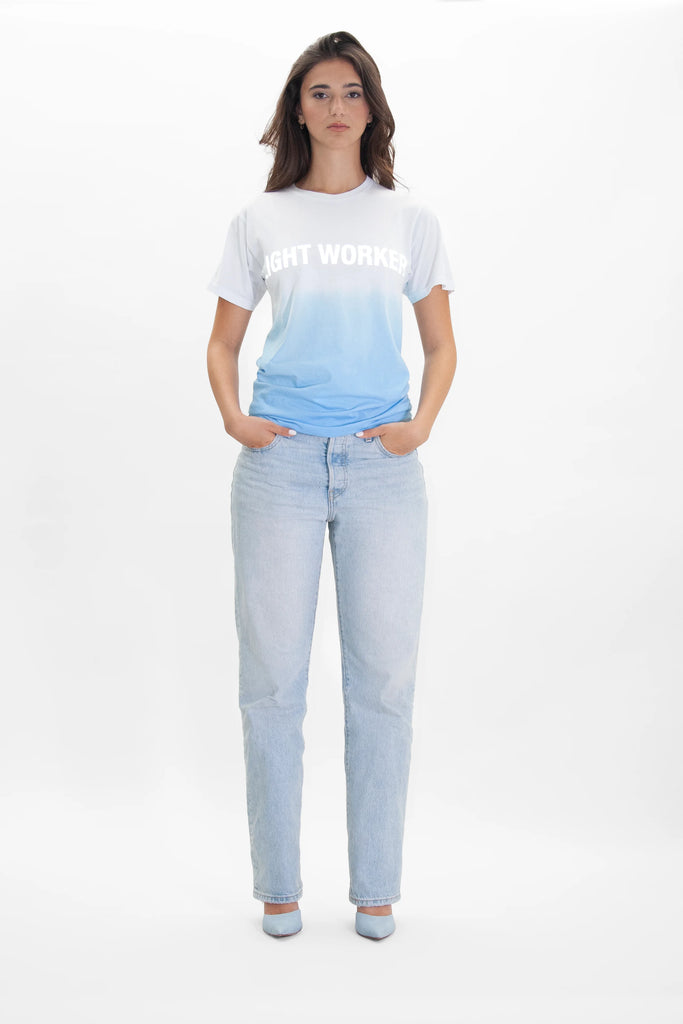 A person stands facing forward, wearing a white and blue "Light Worker Tee in Atmosphere" from GFLApparel with a reflective print. They have on light blue jeans and light blue shoes, all set against a plain white background. Hands are in pockets.