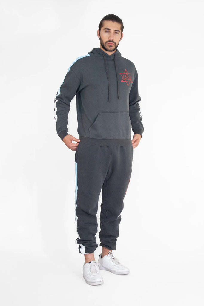 A man with a beard stands wearing a dark GFLApparel Merkaba Hoodie in Space Glow with a star design inspired by sacred geometry, matching jogger pants, and white sneakers against a plain white background.