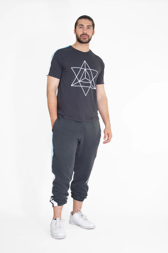 A man with a beard is standing and wearing a black GFLApparel Merkaba Tee in Space Glow, black sweatpants, and white sneakers against a white background.