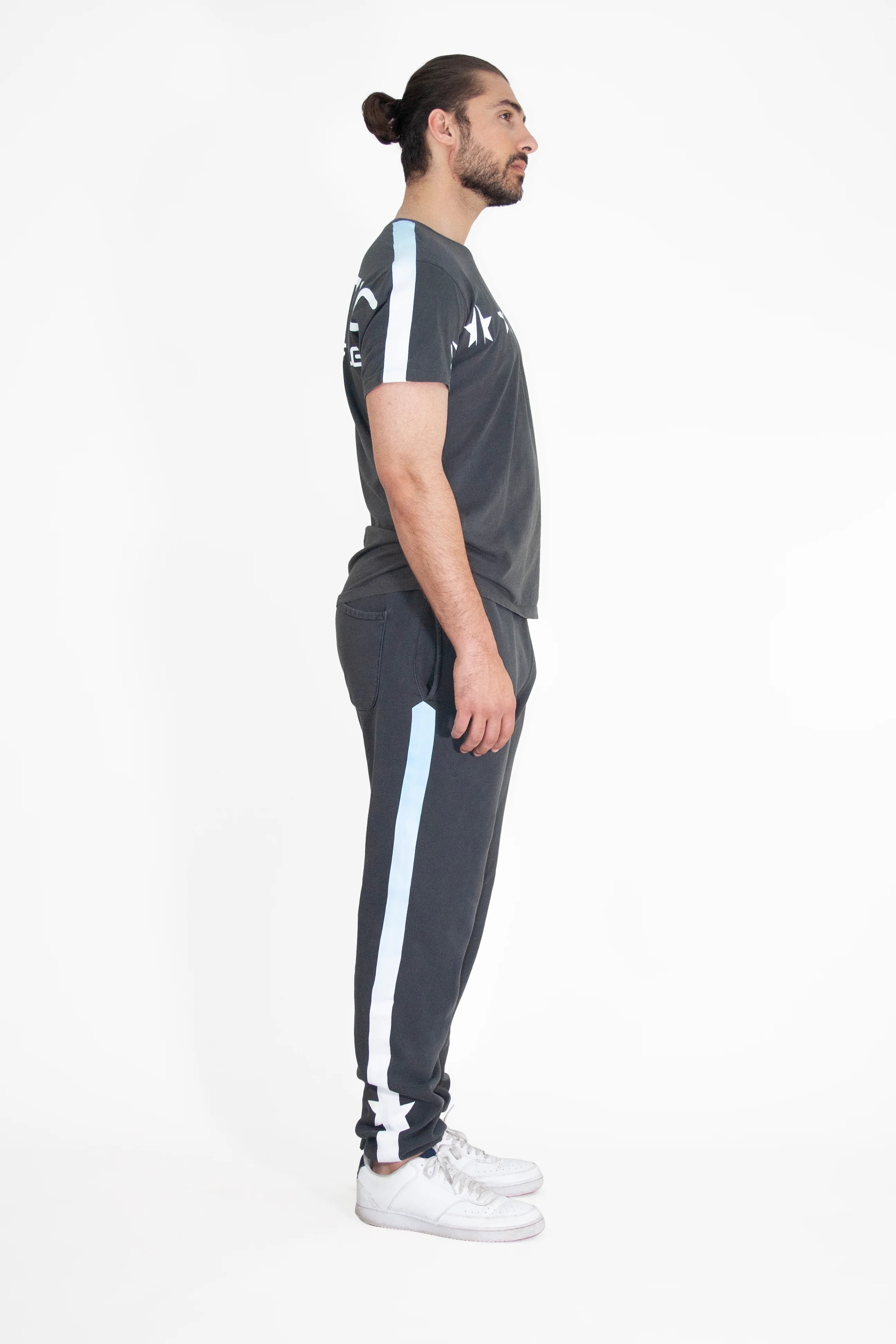 A man with a ponytail is standing in profile, wearing a dark GFLApparel GFL Stars Tee in Space Glow and matching dark pants with light blue stripes. His relaxed fit attire is completed with white sneakers, all set against a plain white background.