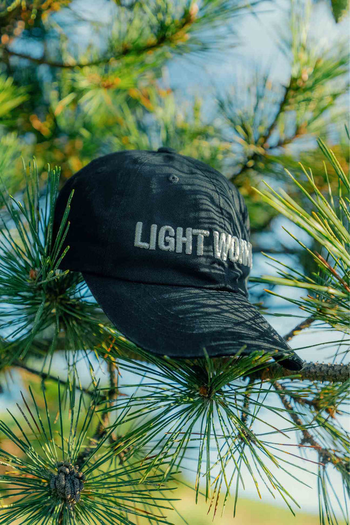 A must-have Light Worker Baseball Cap from GFLApparel with the words "LIGHT WORK" embroidered on it is placed on the branches of a pine tree, making it a staple piece for any Light Worker.